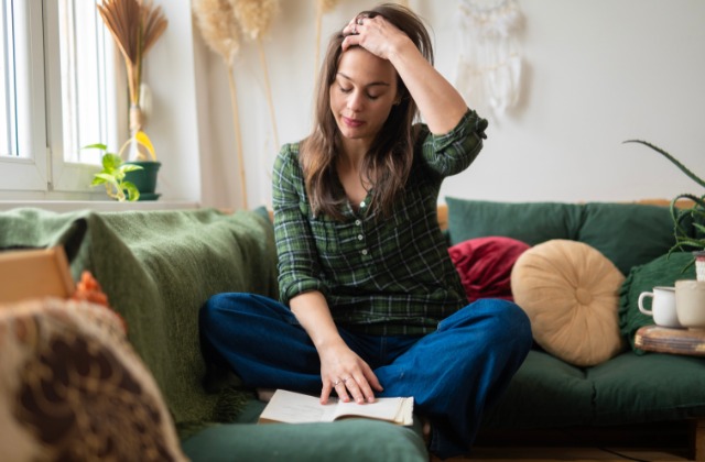A young college student sits cross-legged on a green couch, holding her head while reading a book in her living room.