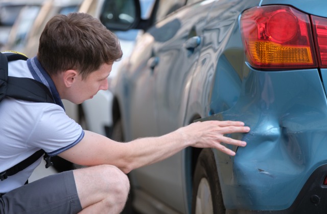 A college student with a backpack examines a dent on the rear panel of his blue car in a parking lot.