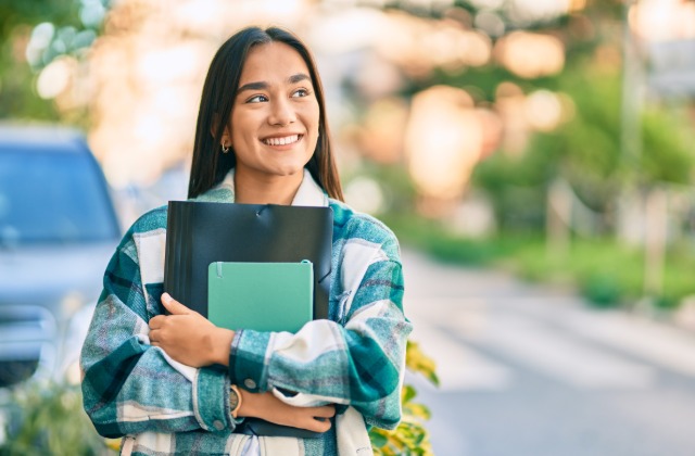 A college-age woman smiling broadly as she poses with a binder and notebook clutched to her chest outside.
