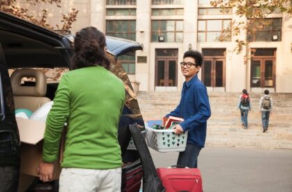 A young man unloads a car with an older adult outside a building, carrying a laundry basket and a suitcase.