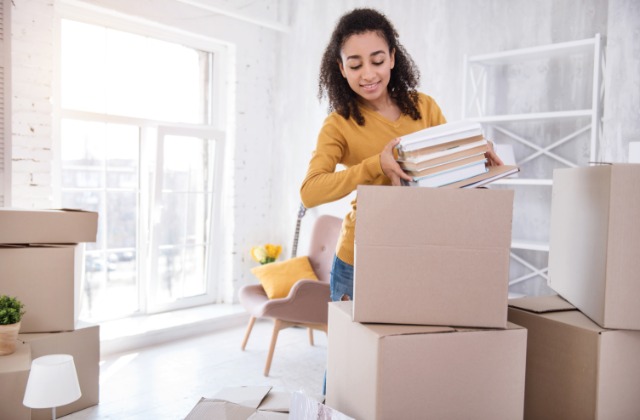 A woman in a yellow shirt is placing a stack of books into a cardboard moving box in a bright, minimalist apartment.