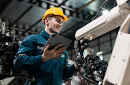 Engineer in a hard hat checks robotic welding arms with a tablet on a factory floor during production.