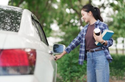 A female student in jeans and a flannel shirt holds books and coffee as she unlocks a white car outdoors.
