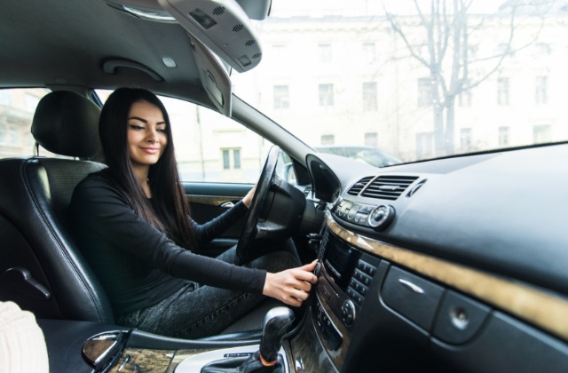 A woman in a black shirt is sitting in the driver’s seat of a car. She’s adjusting some controls on the dashboard.