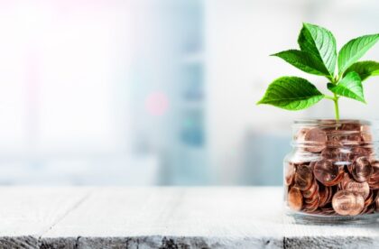 A tiny plant with green leaves is growing out of a glass jar that is filled to the brim with pennies.