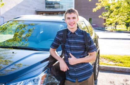 A young man in a striped button-up and a backpack on, leaning on his car as he smiles and gives a thumbs up.