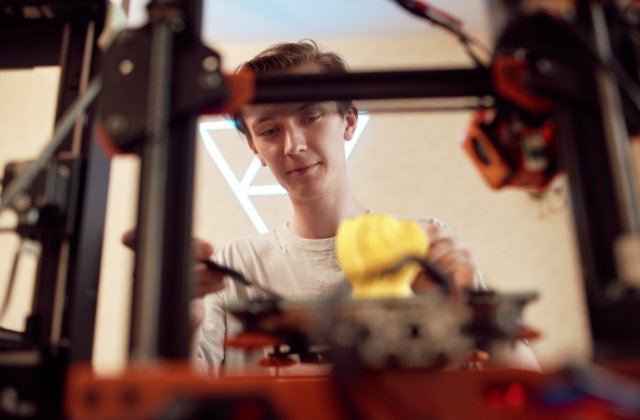 A young man works to pry off a yellow part from a 3D printer's bed. The man's face is framed by the printer's bars.