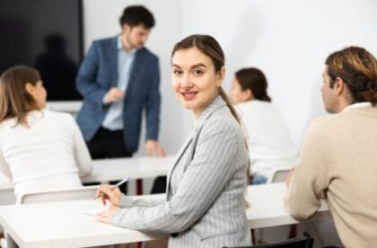 Professional woman writing in notebook during meeting, looking at camera with coworkers and speaker behind her.