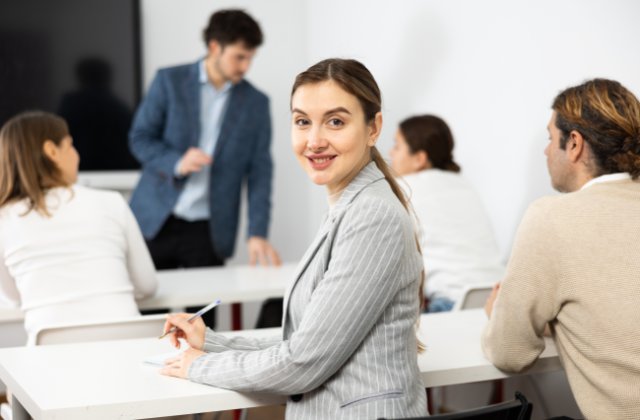Professional woman writing in notebook during meeting, looking at camera with coworkers and speaker behind her.