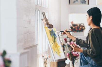 A young woman painting on a canvas held on an artist's easel. She is in front of a window that sunlight is streaming in from.