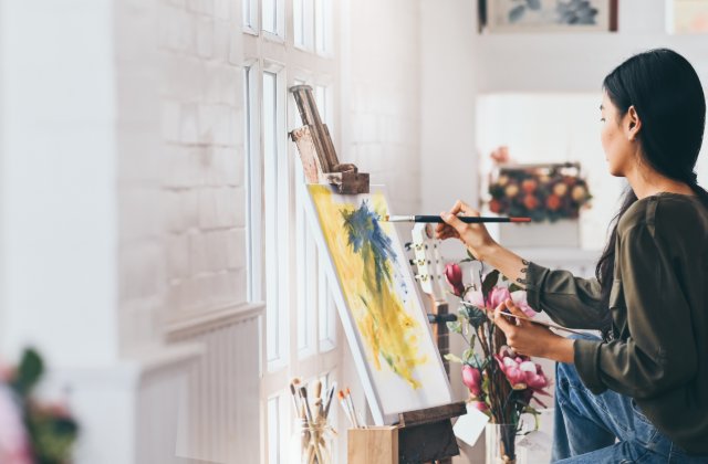 A young woman painting on a canvas held on an artist's easel. She is in front of a window that sunlight is streaming in from.
