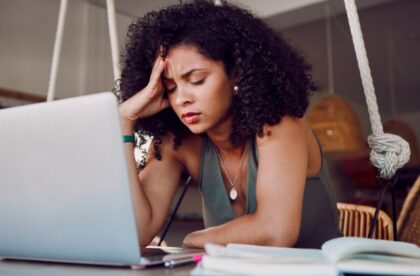 A young woman sitting alone at a desk with an open book and laptop in front of her, while resting her hand on her head.