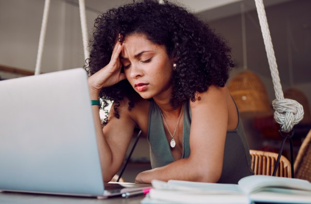 A young woman sitting alone at a desk with an open book and laptop in front of her, while resting her hand on her head.