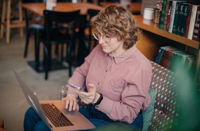 A young woman sits in a comfy chair in front of a bookshelf with a laptop on her lap. She holds her phone in one hand.