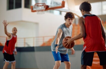 Two boys play basketball indoors with a hoop and gym seating in the background. One dribbles while the other defends,