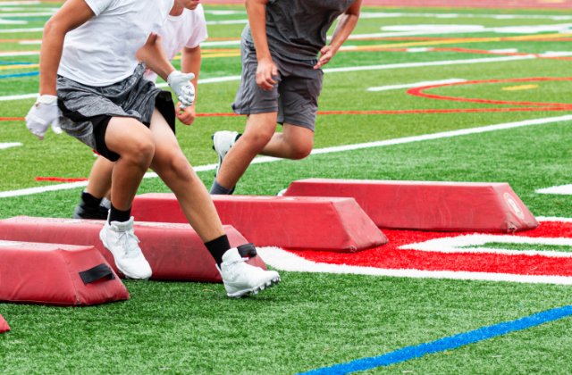 Three young boys navigating red barriers as part of an athletic practice drill on a turf football field.
