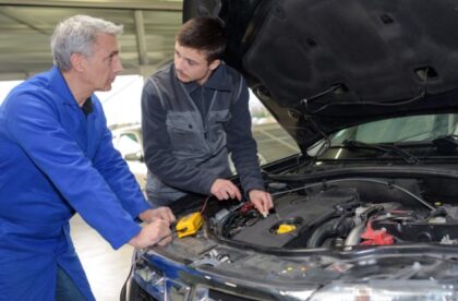 A college-age person and an older mechanic leaning over an open car hood. The student handles wires under the hood.