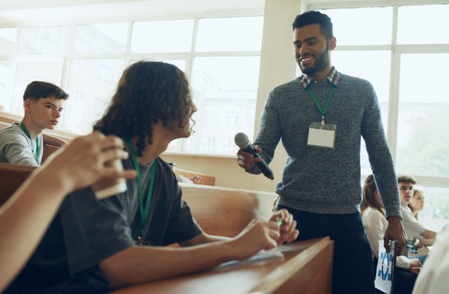 A man stands between rows of students in a lecture hall and points a microphone at one of them as he smiles.