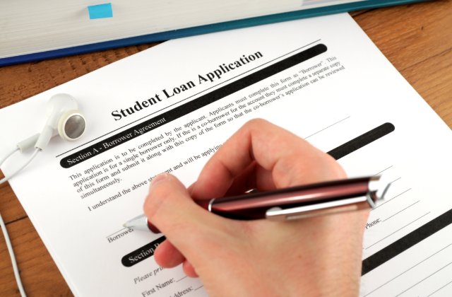 A person fills out a student loan application with a red pen on a wooden desk beside a book and earbuds.