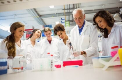 A group of people wearing white lab coats watching an older man use a pipette at a laboratory table.
