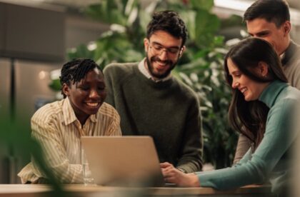 A close-up of four people standing together and smiling while looking at a laptop set up on an office desk.