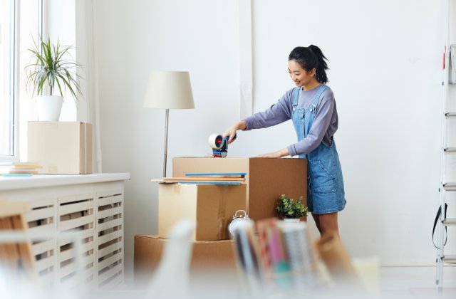 Multiple cardboard boxes are stacked in a room with a dresser and white walls. A woman tapes a box shut.