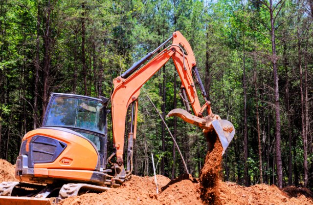 An orange excavator is positioned on a mound of dirt and is releasing dirt onto the ground. Tall trees fill the background.