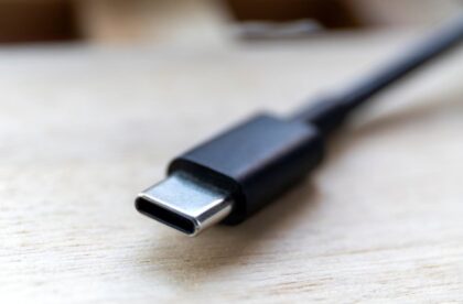 An extremely close-up view shows a black USB-C charging cord lying flat on a wooden surface. The end is metal.