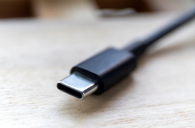 An extremely close-up view shows a black USB-C charging cord lying flat on a wooden surface. The end is metal.