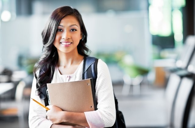 A student with long, curled hair holds a notebook and a pencil. She wears a backpack in a bright, blurred indoor space.