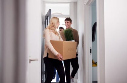 Friends move into a bright home, carrying a cardboard box and tall green plant through the open front door.