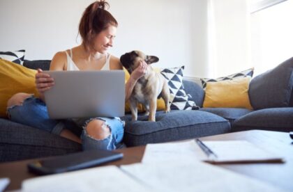 A college student pets a small dog while using a laptop on a couch, with study materials on the table.