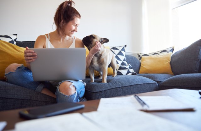 A college student pets a small dog while using a laptop on a couch, with study materials on the table.
