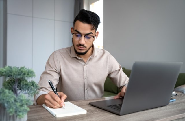 A man wearing glasses writing in a notebook at his desk beside an open laptop and a small potted plant.