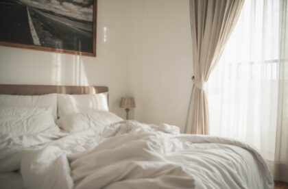 A cozy master bedroom with white linens and floor-to-ceiling curtains in a beige color. The bed is unmade.