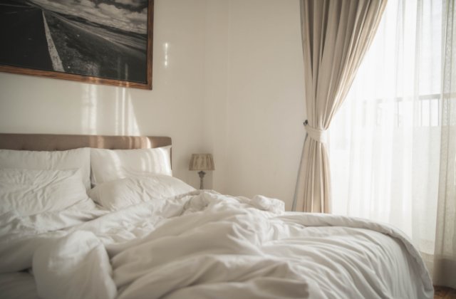 A cozy master bedroom with white linens and floor-to-ceiling curtains in a beige color. The bed is unmade.