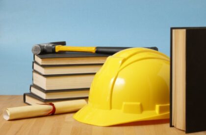 A yellow hard hat resting beside a hammer, stacked books, and a rolled diploma tied with a blue ribbon.