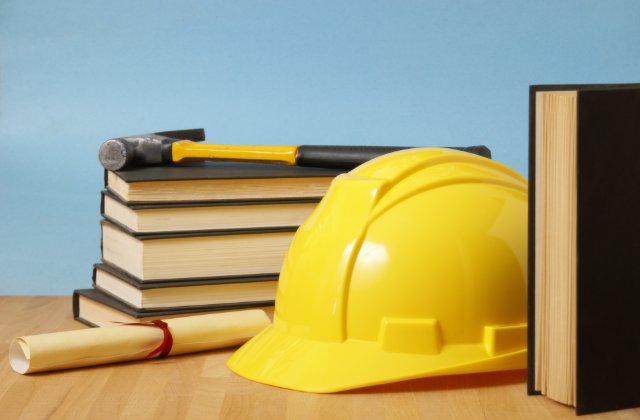 A yellow hard hat resting beside a hammer, stacked books, and a rolled diploma tied with a blue ribbon.