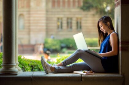 A college student using a laptop while seated on a stone ledge near columns, with a notebook and eyeglasses beside her.