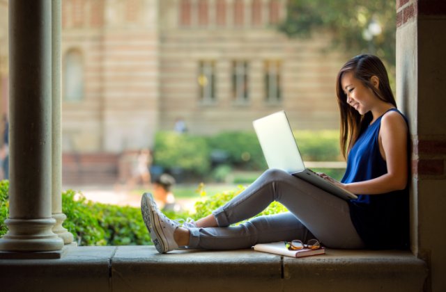 A college student using a laptop while seated on a stone ledge near columns, with a notebook and eyeglasses beside her.