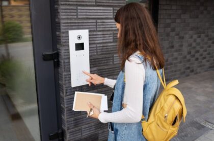 Woman with a yellow backpack enters a security code on a building keypad while holding an envelope and keys.