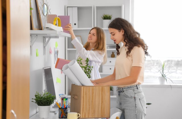 Two college students unpack decorations and supplies while organizing shelves in a bright, modern room.