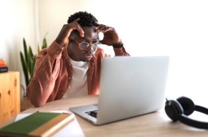 A male college student sits at a desk with his hands on the side of his forehead, looking at an open laptop.