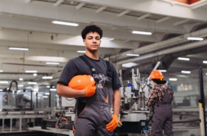A young factory worker holding an orange hard hat while another worker operates industrial machinery in the background.