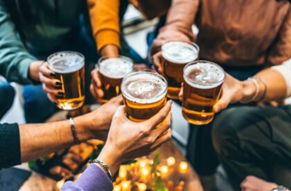 Several people raise glasses of beer together during a casual social gathering, with hands meeting in the center.