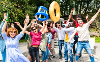 A group of people outdoors holding a boombox and bottles while smiling, cheering, and standing together in sunlight.