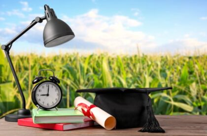 A black graduation cap, rolled diploma, lamp, clock, and books resting on a wooden table before a green field.