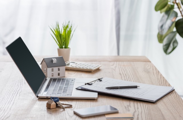 A close-up of a desk with a laptop, model house, keys, contract, calculator, smartphone, and potted plant.