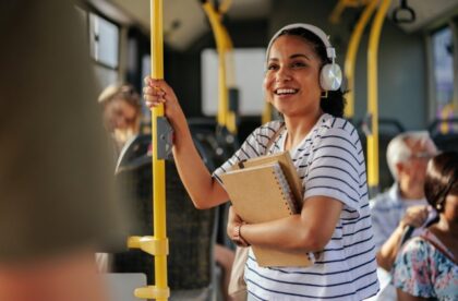A woman wearing headphones holding notebooks and a yellow pole while standing among passengers on a bus.