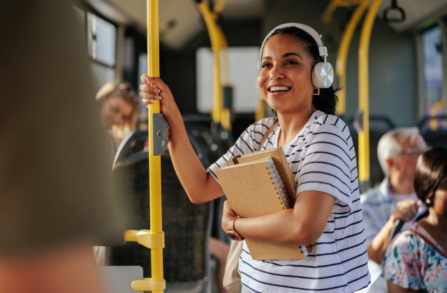 A woman wearing headphones holding notebooks and a yellow pole while standing among passengers on a bus.